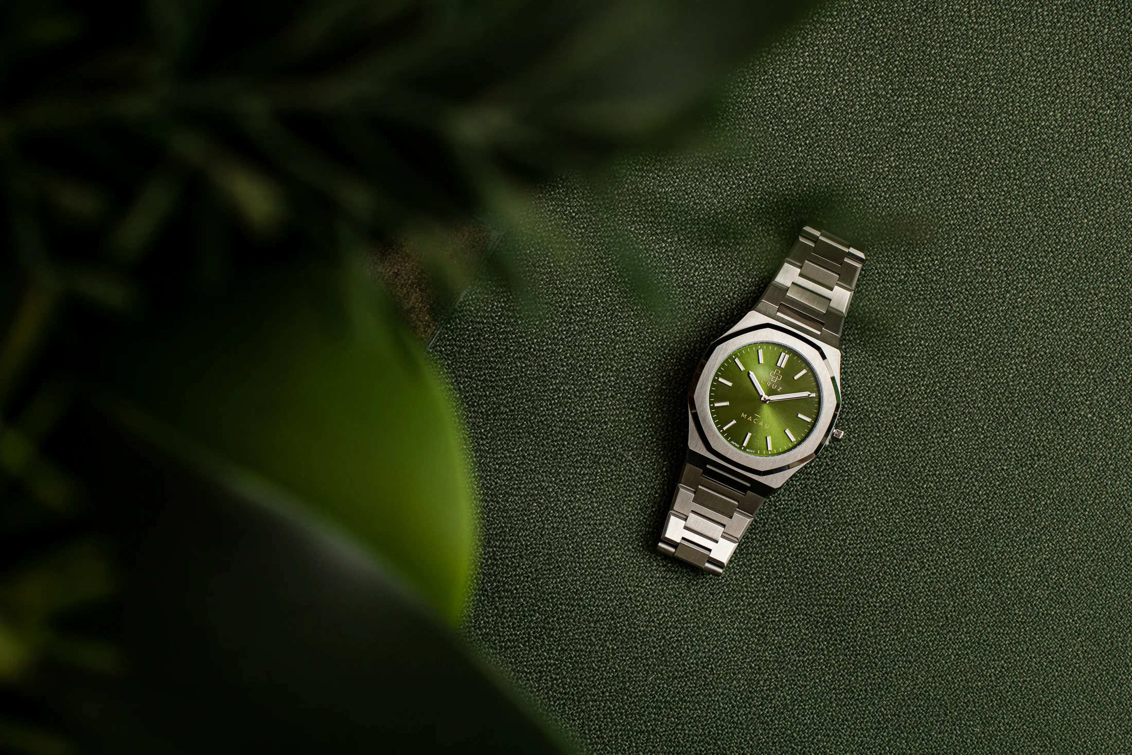 Relógio Cruz Watches Macau de mostrador verde, sobre tecido verde-escuro, com pulseira prateada e caixa octogonal.